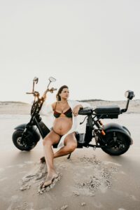 A pregnant woman sits on the beach with a motorcycle, embodying leisure and style.